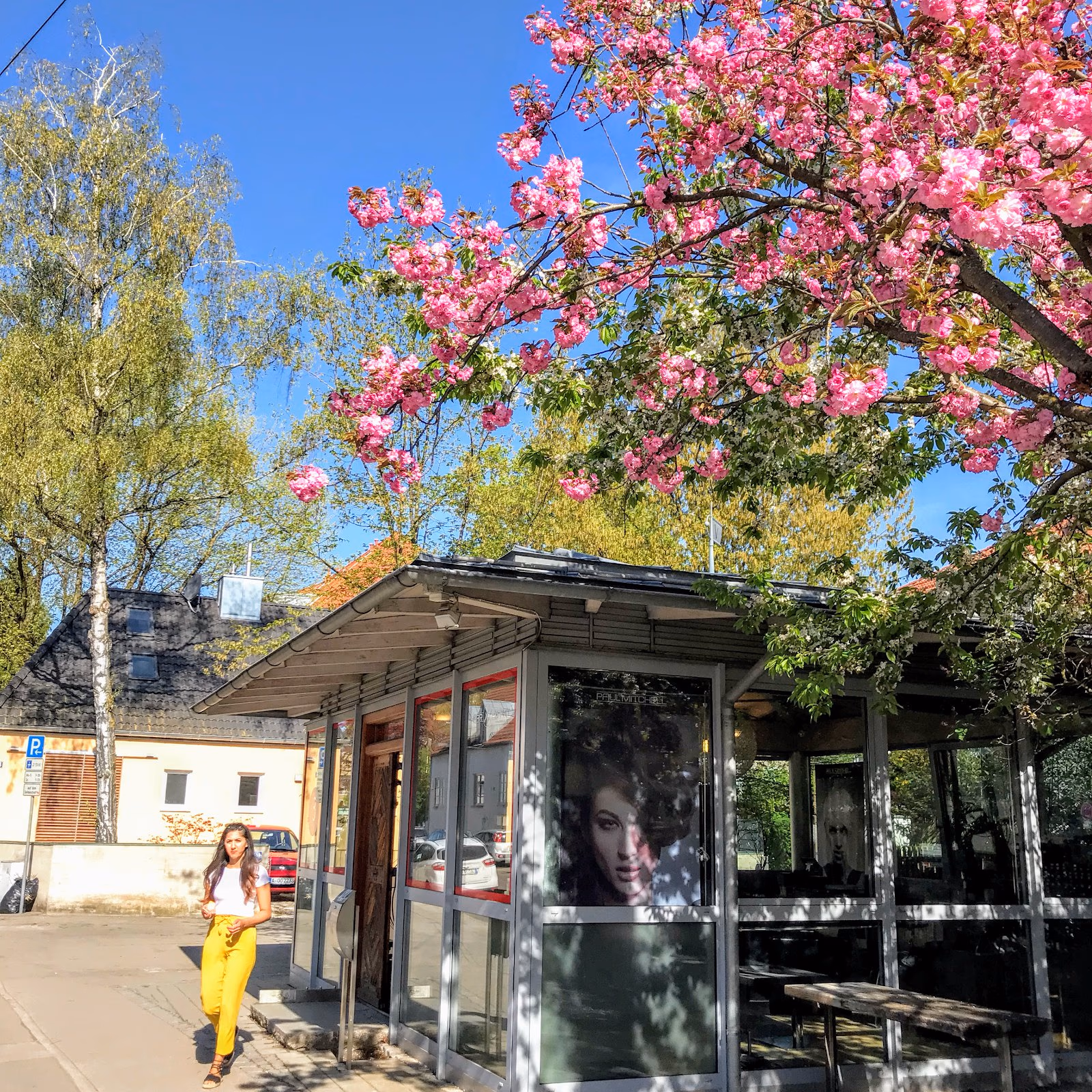 Der Salon Haar Exakt in Augsburg-Pfersee von außen, ein Glaspavillon mit Holzdach unter blühenden Bäumen